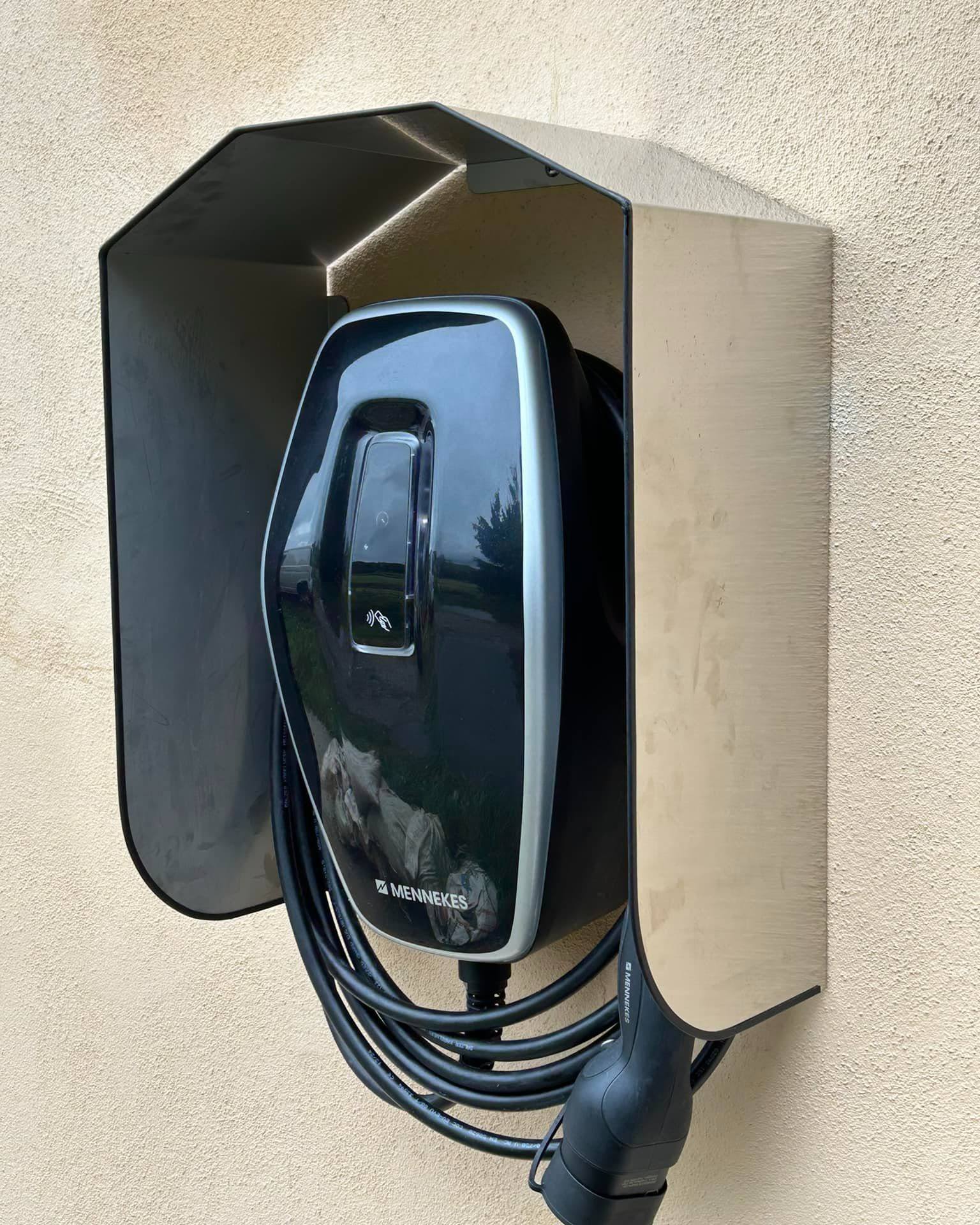 Mennekes electric vehicle charger installed on an exterior wall, protected by a sleek stainless steel weatherproof cover. The charging cable is neatly coiled below the unit.